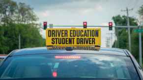 A car driving with a yellow "DRIVER EDUCATION STUDENT DRIVER" in all caps sign image is a shot of the car's roof in close rear view