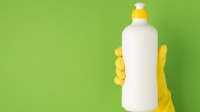 Yellow gloved hand holding a white bottle of dish soap in front of a green background.