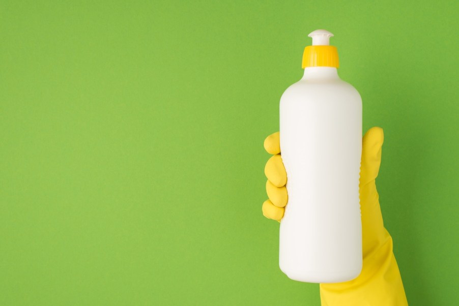 Yellow gloved hand holding a white bottle of dish soap in front of a green background.