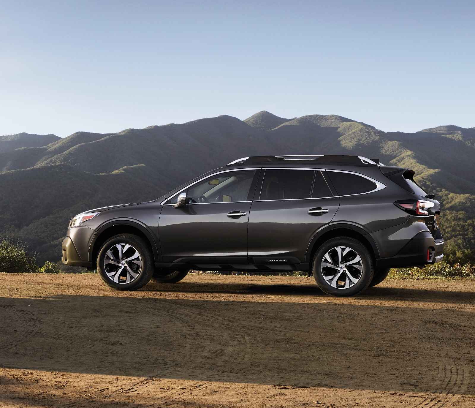 A dark grey 2022 Subaru Outback Touring parked in full left profile view on a desert ground with mountains in background
