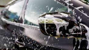 A car wash features a black hatchback covered in suds and soap.
