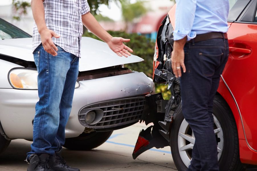 Two drivers confront one another during a road rage incident, a car crash visible in the background.