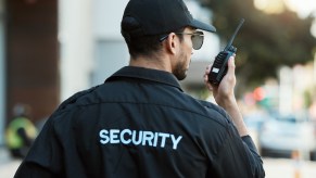 Man in security jacket, hat and sunglasses speaks on a walkie talkie, a city street visible in the background.