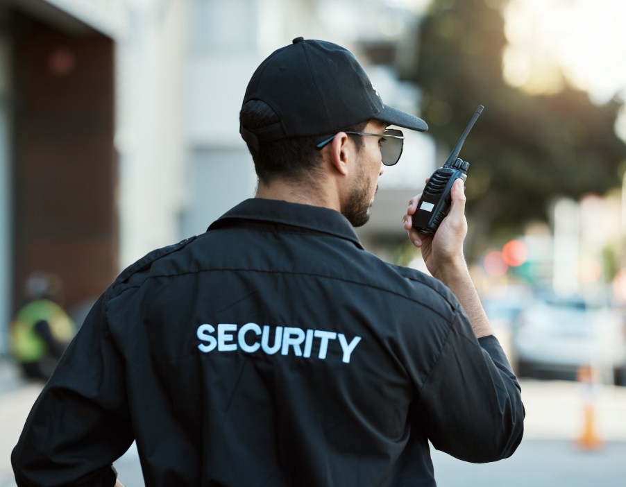 Man in security jacket, hat and sunglasses speaks on a walkie talkie, a city street visible in the background.