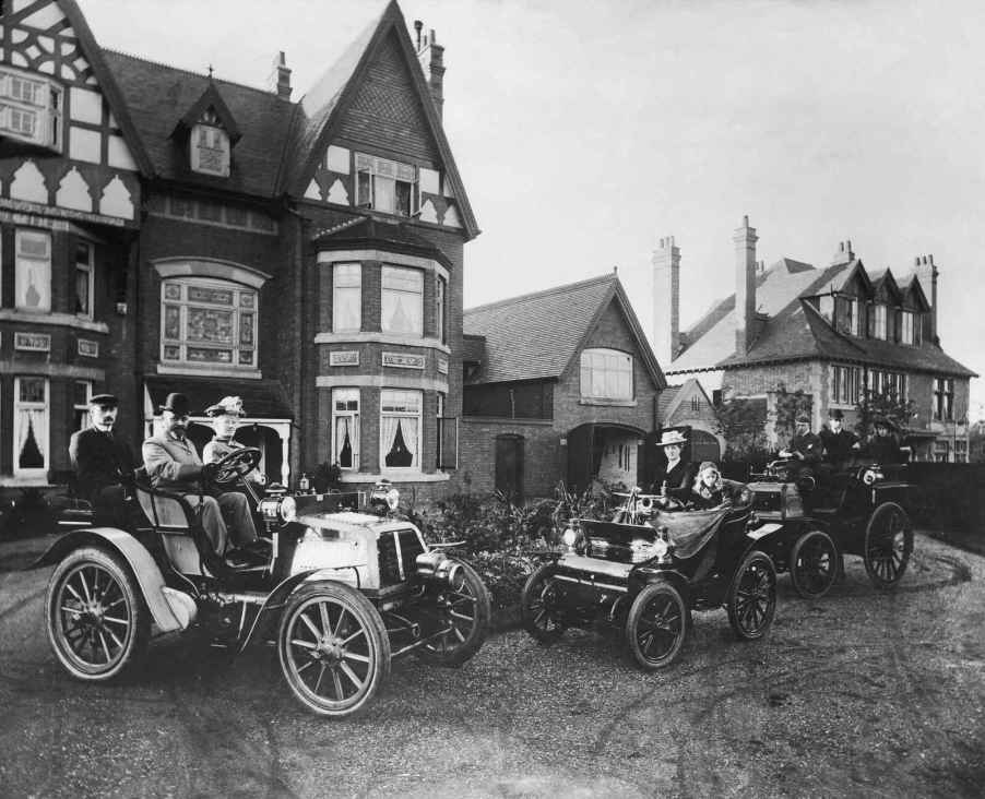 A 1910 photo of several groups of people sitting in early cars in front of a large brick estate. Depiction of earl cars and their license plates in the U.S.