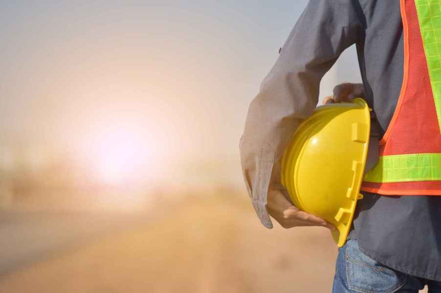 A road worker shown from shoulders down holding a hard hat against his left hip