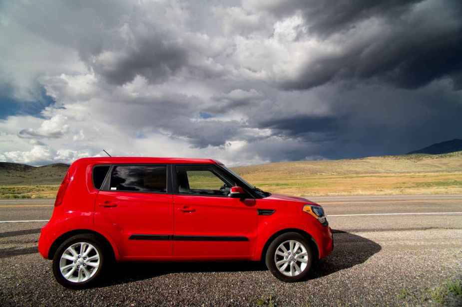 A red Kia Soul parked on the side of the road with storm clouds in the distance