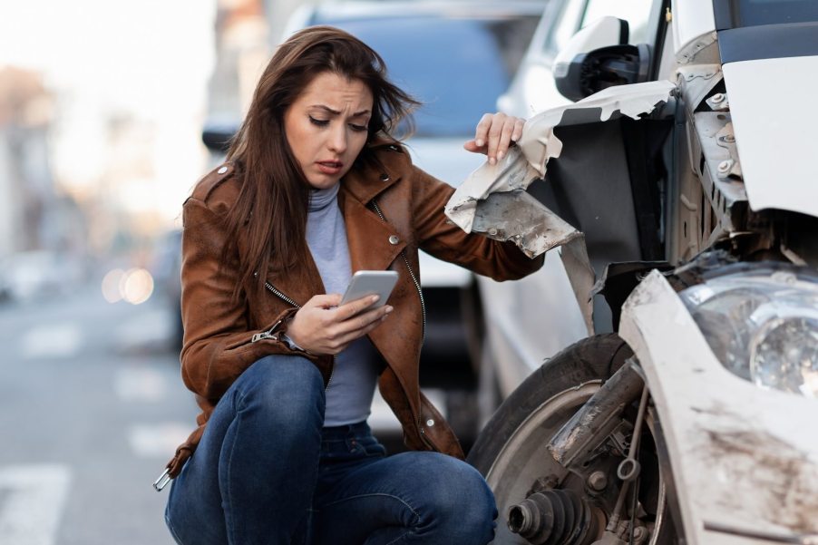 Young woman crouches by her crashed car to take a photo with her cellphone