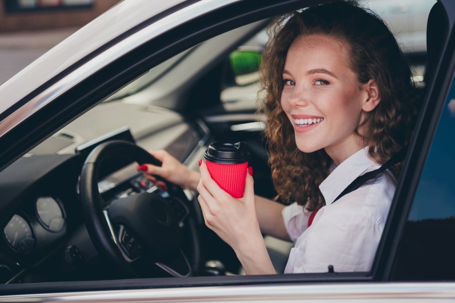 A woman at the wheel of a car holds up a to-go cup of tea