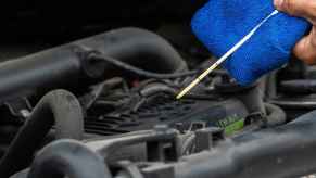 A man's hand holding blue towel and engine oil dipstick in front of open hood in very close view