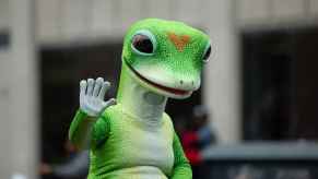 Geico insurance company gecko mascot waving on top of a car during a parade in close waist-up view