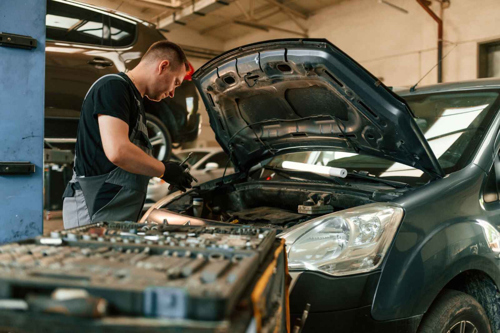 An automotive technician leaning over a vehicle using a wrench, with a cart full of tools at the ready