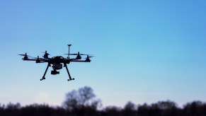 Remote controlled drone with a camera visible in the blue sky, over a tree line