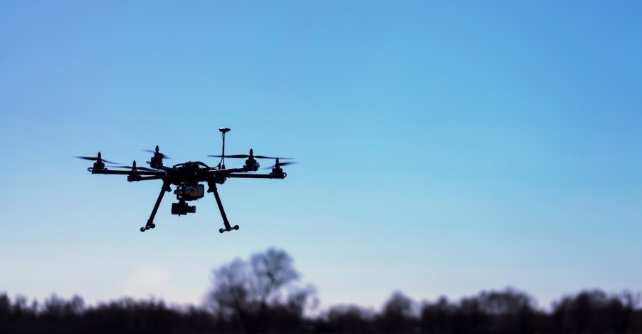 Remote controlled drone with a camera visible in the blue sky, over a tree line