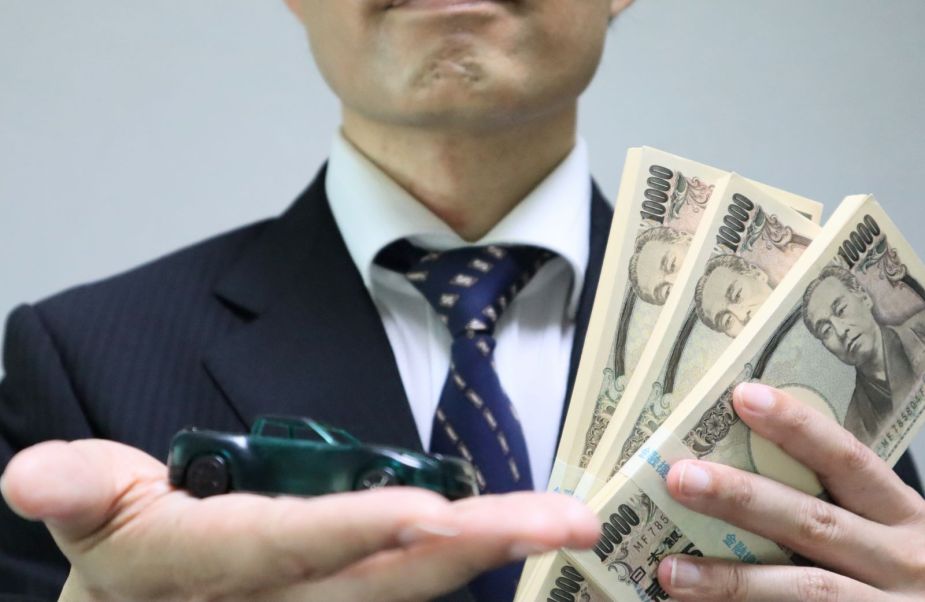 A man holding up a toy car and stacks of money, representing a scam involving cars