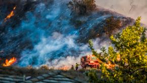 Two firefighters stand next to the LA County "Fire Dozer" while a hillside burns in the background.