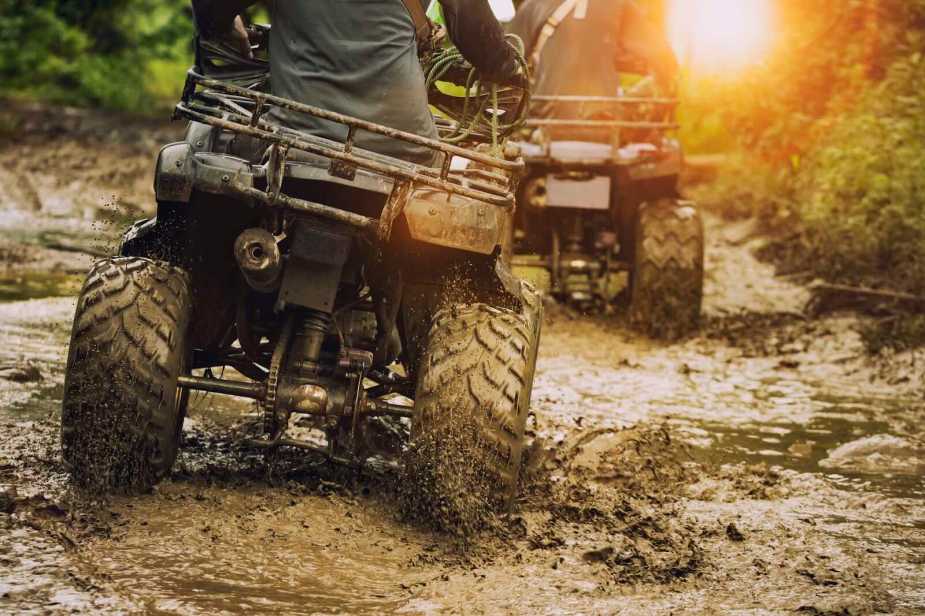 Two ATV riders crossing a dirt road, the setting sun in the background.