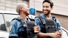 Louisiana police officers standing by their cruiser, drinking coffee, and laughing about their traffic bingo game cards.