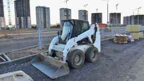 White skid-steer vehicle parked at a construction site, buildings in the background.