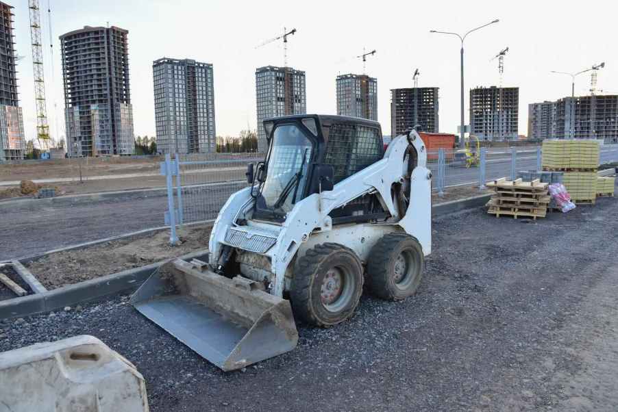 White skid-steer vehicle parked at a construction site, buildings in the background.