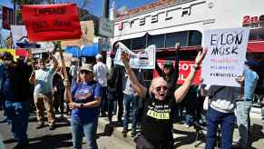 Rows of protestors shouting outside a Tesla dealership, holding signs against CEO Elon Musk and the trump administration
