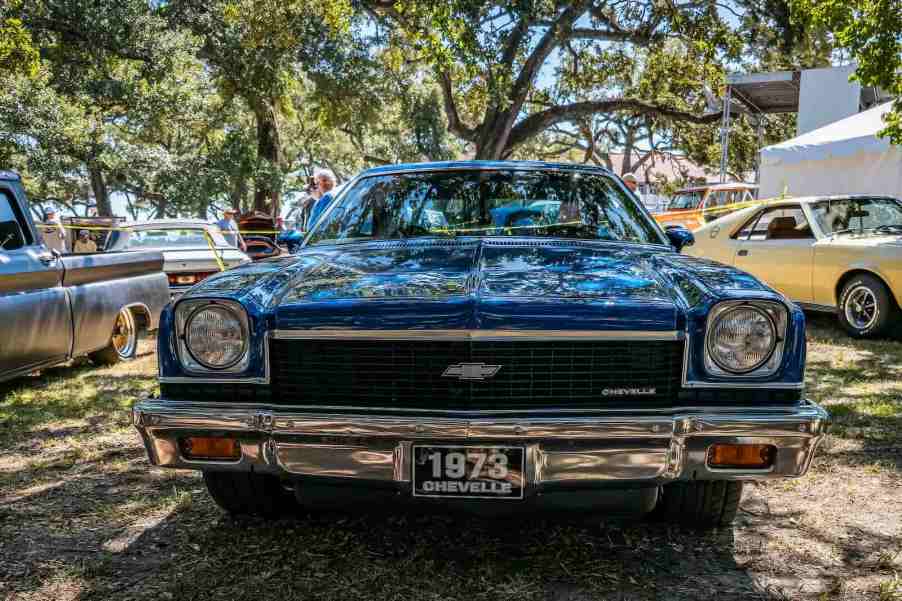 Blue Chevrolet Chevelle muscle car parked under trees at an auto show.