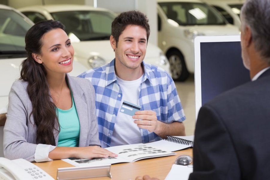 Car dealership salesman signing papers with vehicle buyers.