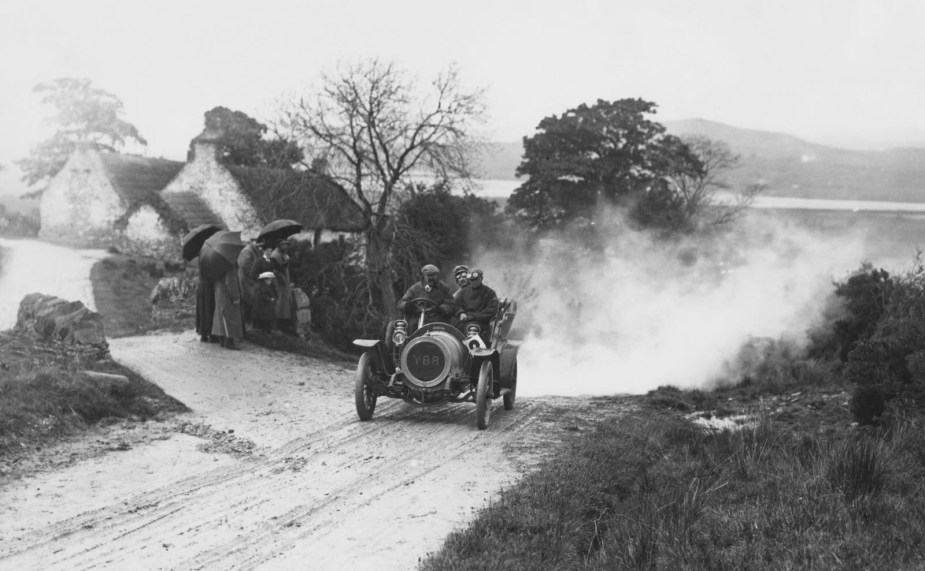 Race car blasts down a dirt road between farmhouses, spectators look on.
