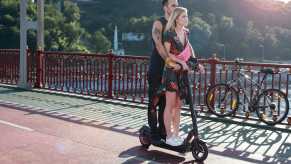 Man and woman ride an electric scooter over a Florida bridge, water visible in the background.