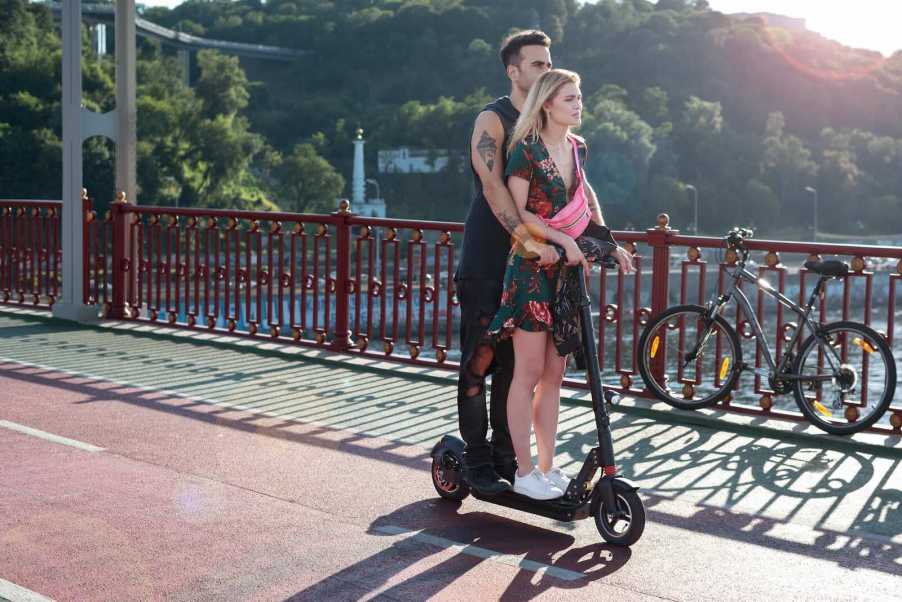 Man and woman ride an electric scooter over a Florida bridge, water visible in the background.