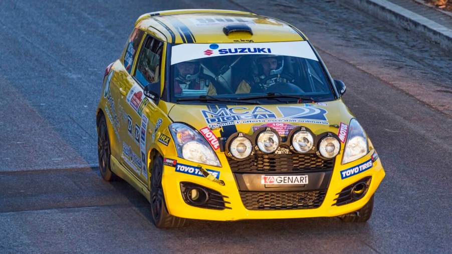 Yellow rally spec Suzuki car with extra headlights racing on a track.