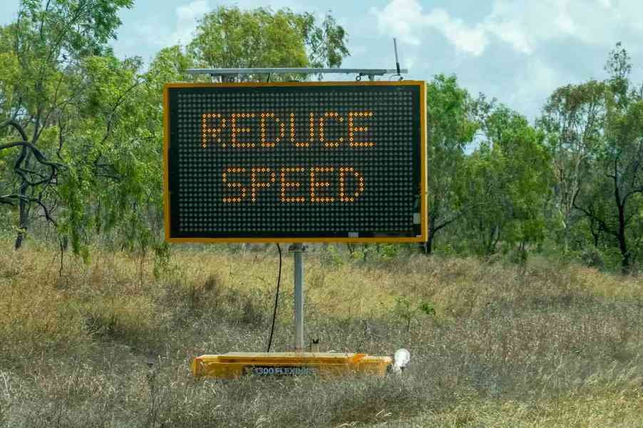 LED sign warning drivers "reduce speed" ahead of MPH limit, trees visible in the background.