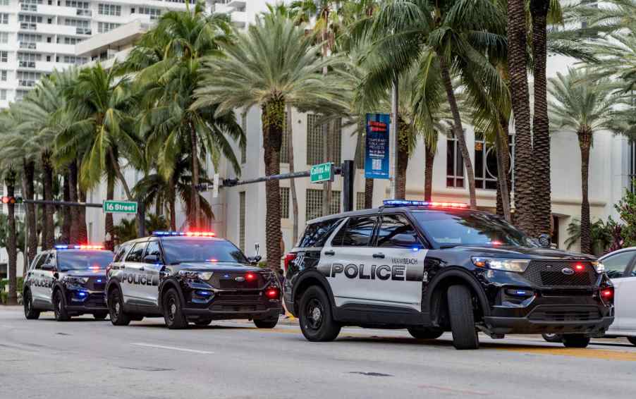 Several SUV police cruisers pulled over facing the viewer with roof lights on during the day