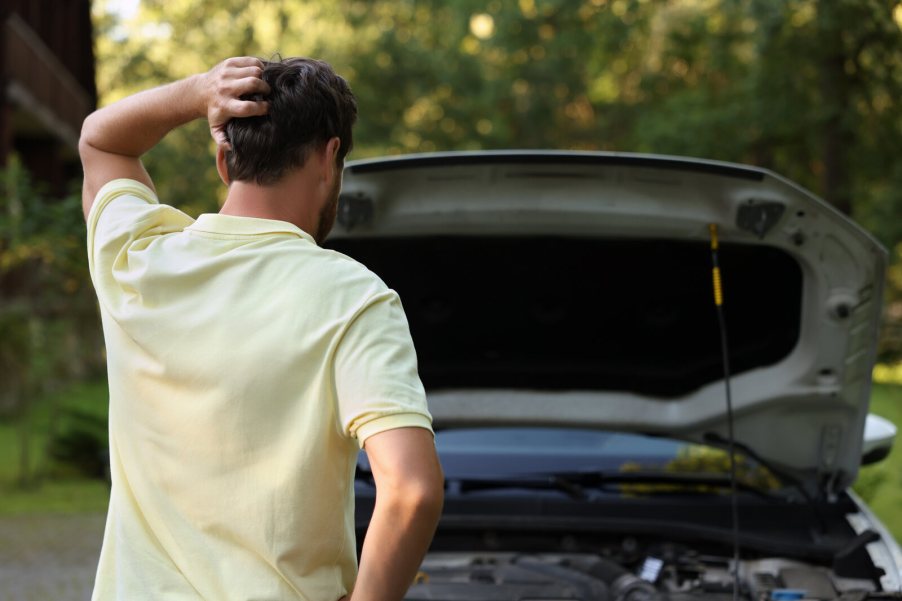Driver standing in front of open car hood holding left hand to back of head facing away from viewer
