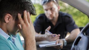 Driver with their head in their hands, a cop in the background during a police traffic stop for a suspended license because of unpaid child support
