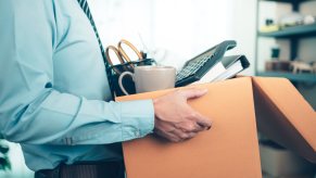 A man in a shirt and tie holding a cardboard box full of office supplies quitting job