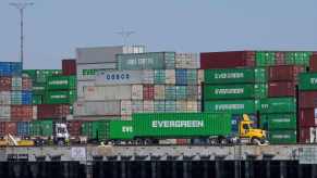 A semi-truck loaded with a shipping container parked in front of a stack of shipping containers at the Port of Los Angeles