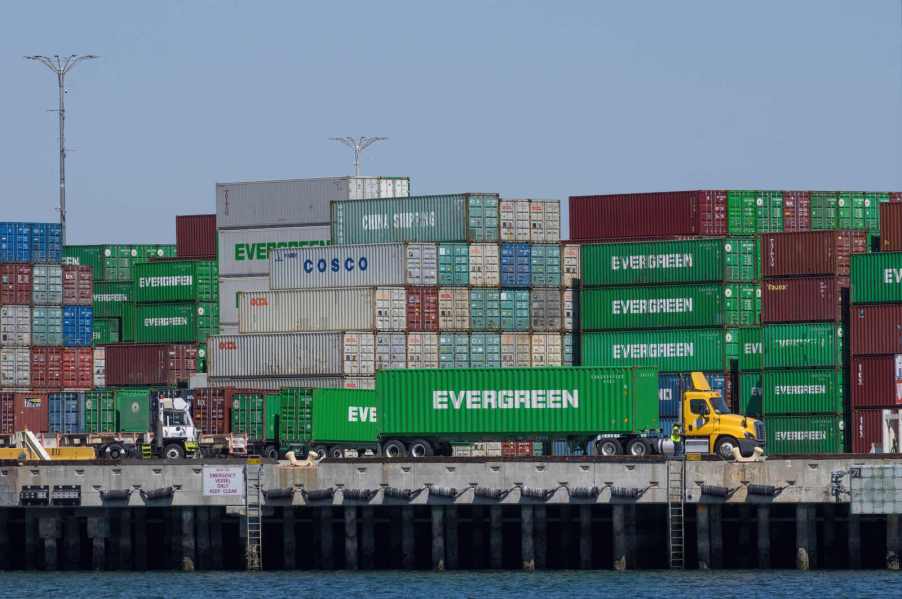 A semi-truck loaded with a shipping container parked in front of a stack of shipping containers at the Port of Los Angeles