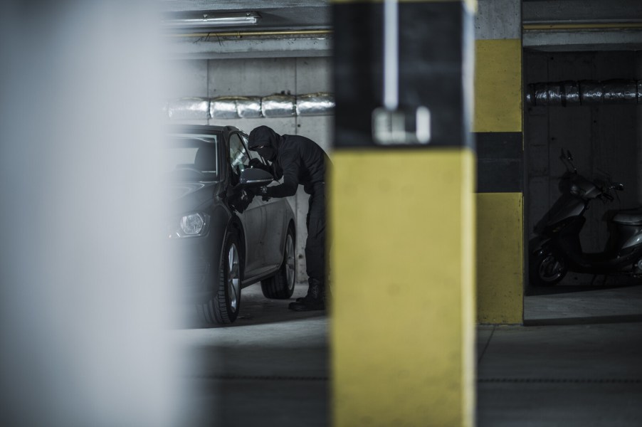 Man dressed in all black and black mask looks into car window parked in dark parking garage in far view