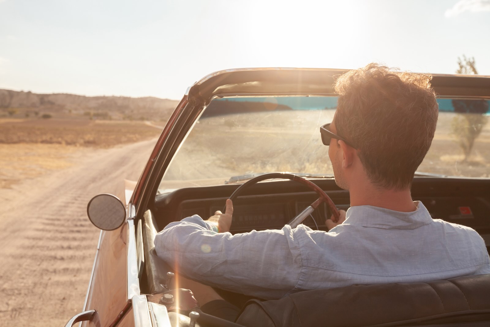 Man wearing old money style clothes while driving a classic convertible on a dirt road.