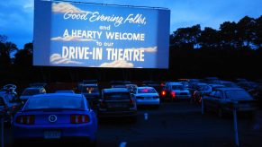 Rows of cars at a drive-in movie theater at dusk, trees in the background.