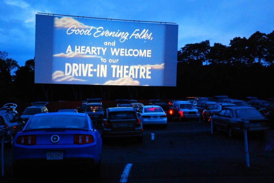 Rows of cars at a drive-in movie theater at dusk, trees in the background.