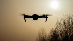 A police drone hovers over a stop sign to monitor and ticket drivers, trees visible in the background.