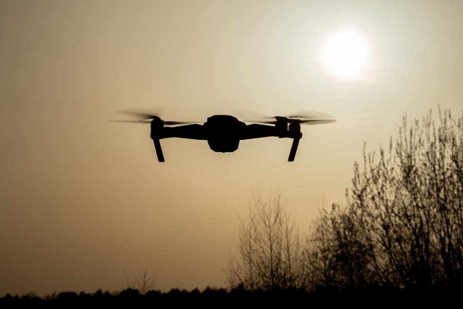 A police drone hovers over a stop sign to monitor and ticket drivers, trees visible in the background.