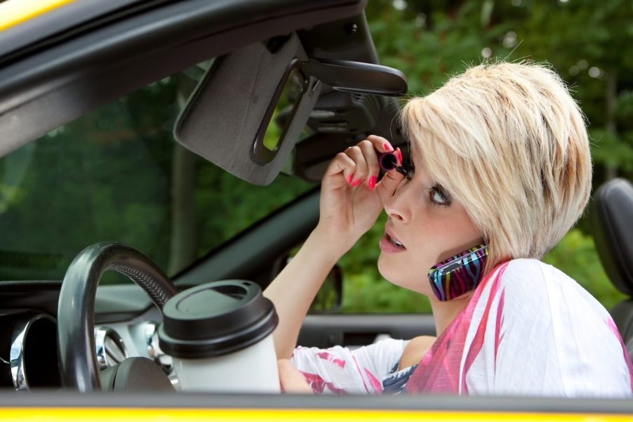 Gen Z driver applying makeup and drinking coffee while behind convertible wheel.