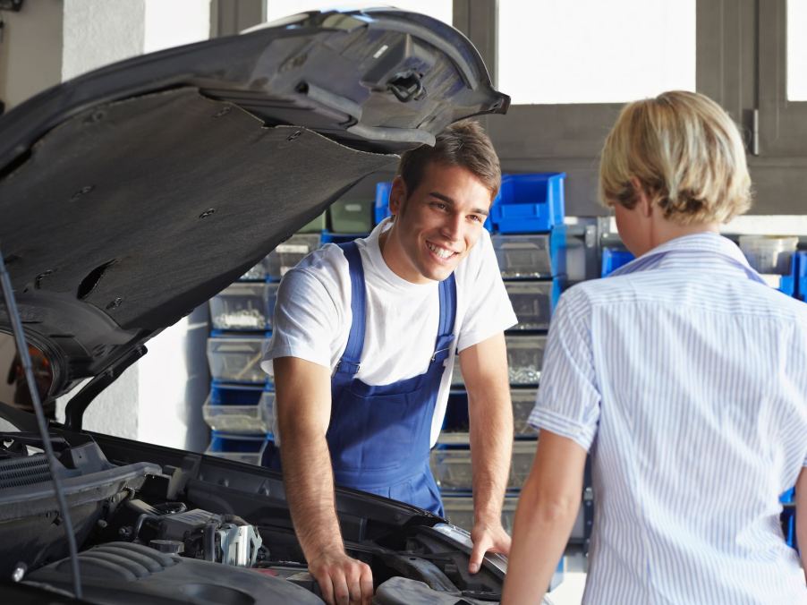 Smiling mechanic talks to female marine over open car hood while attempting to con her.