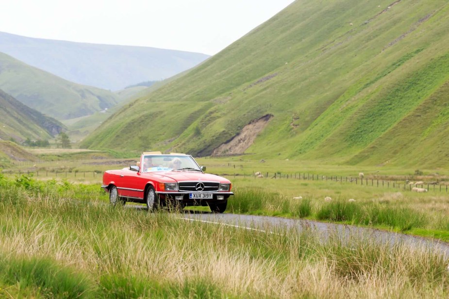 Red Mercedes SL roadster driving through the countryside, mountains visible in the background.