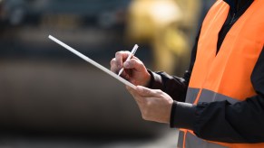 Man in orange neon vest holds a clipboard at a car auction.