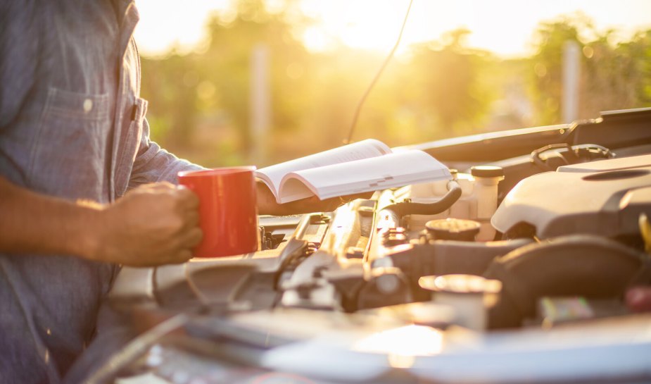 A man holding a coffee cup and reading a manual above an open car engine bay