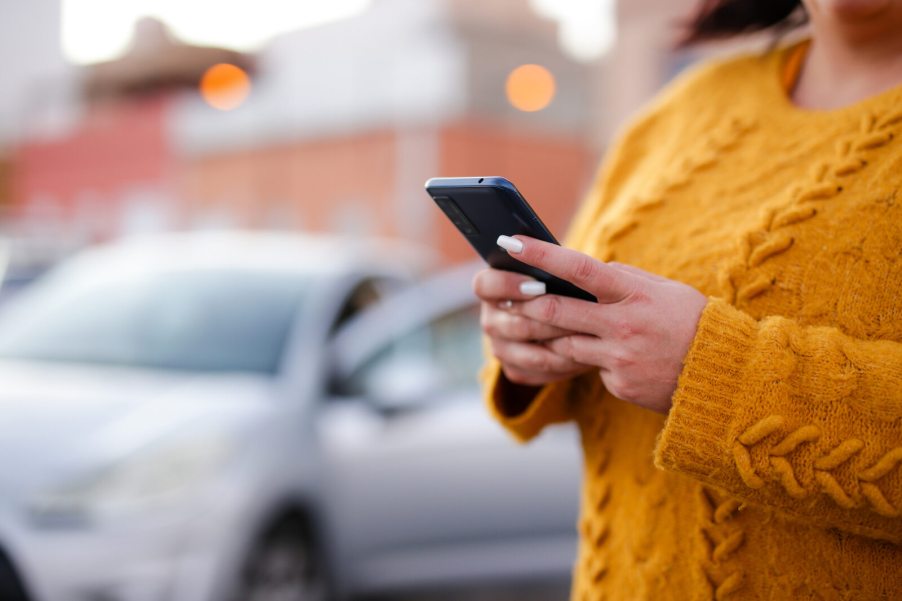 A woman holding a cell phone with a silver car in the background view from shoulder down